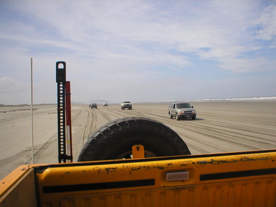 Yellow pickup truck bed with spare tire and hi-lift jack, driving on a sandy beach with other vehicles