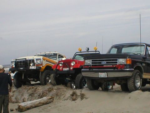 1978 Ford Bronco - Three off-road vehicles are parked on a sandy dune, with a log in the foreground.