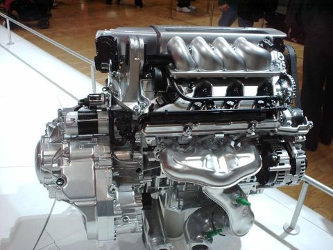 1997 Ford Taurus - Rear three-quarter view of a transverse V8 engine bolted to an aluminum transaxle, displayed on a stand behind a chrome rail at an indoor a…