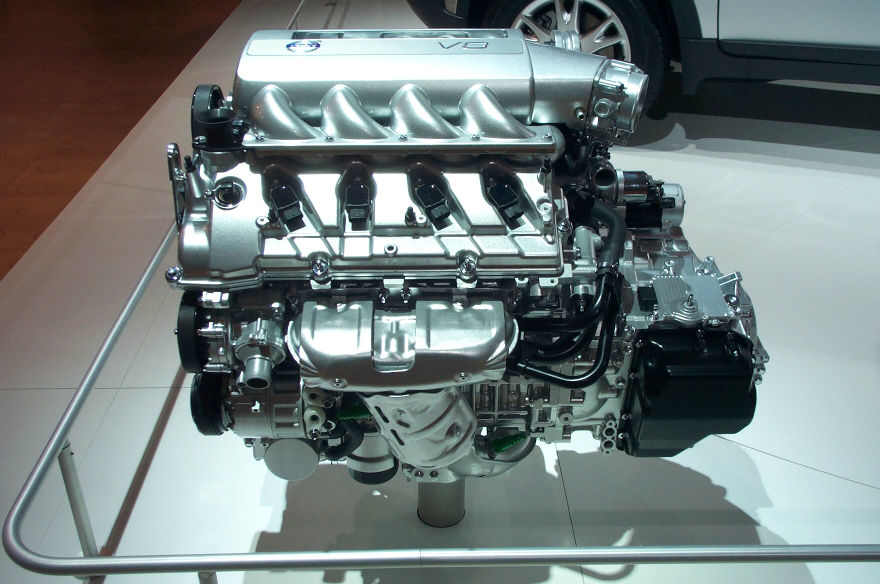Silver V8 engine on display stand, with a white vehicle in the background
