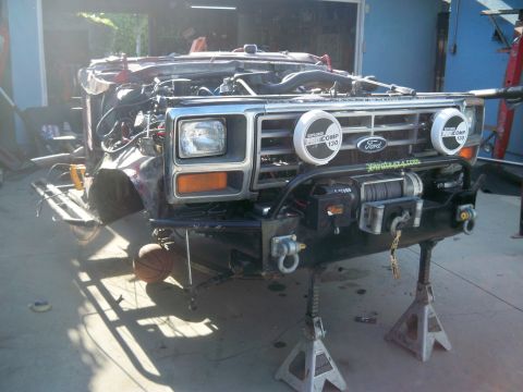 1985 Ford Bronco - The front of a disassembled Ford Bronco, showing its grille, winch, and auxiliary lights, is supported by jack stands.