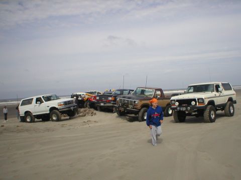 1978 Ford Bronco - A group of Ford Broncos and trucks are parked on a sandy beach under a cloudy sky.