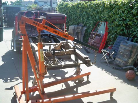1985 Ford Bronco - A red 1985 Ford Bronco chassis is supported by an engine hoist, with its door and fuel tank visible in the background.