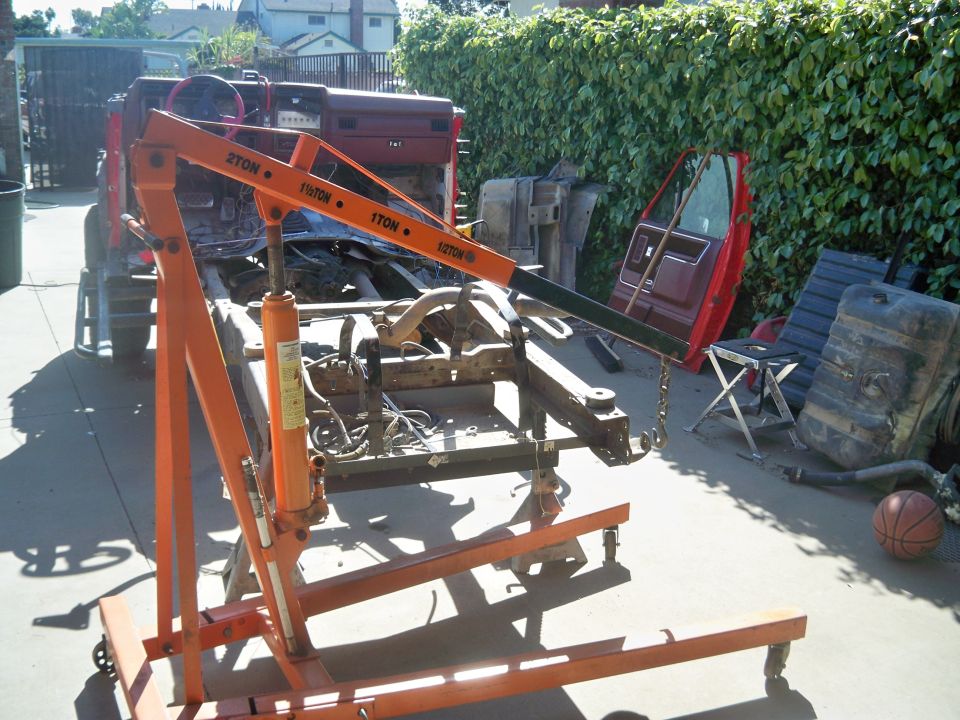 Red 1985 Ford Bronco chassis on engine hoist, door and fuel tank nearby