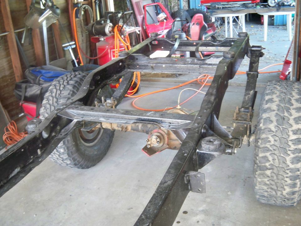 Vehicle frame with leaf springs and rear axle visible. Tires and garage equipment in background