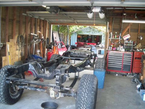 1985 Ford Bronco - The chassis of a vehicle is disassembled and supported by jack stands in a cluttered garage workshop.