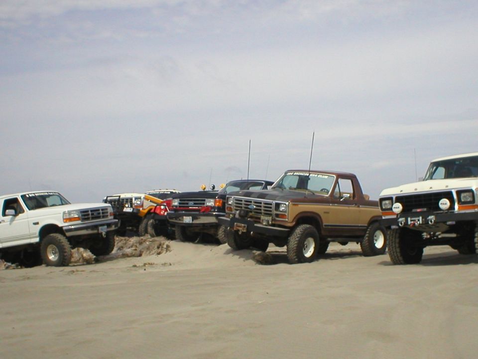 Several 1978 Ford Broncos and trucks parked on a sandy beach under a cloudy sky