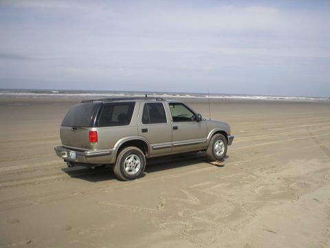 1978 Ford Bronco - A tan SUV sits on a sandy beach, with the ocean waves visible in the distance under a cloudy sky.