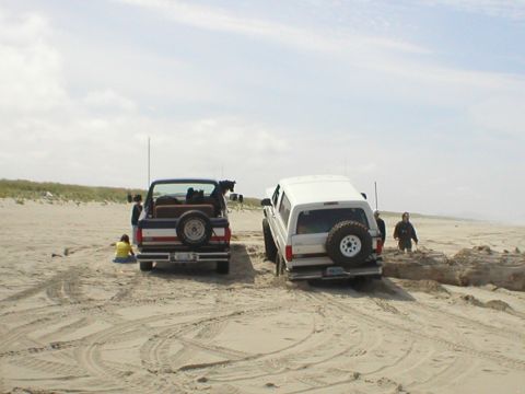 1978 Ford Bronco - Two SUVs are parked on a sandy beach with dunes in the background under a cloudy sky.