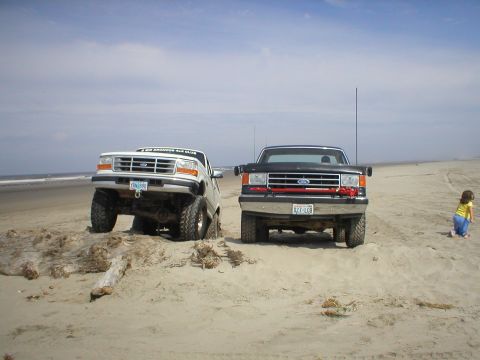 1978 Ford Bronco - Two Ford trucks on a sandy beach with a child in the background.