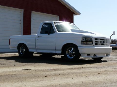 1994 Ford F-150 - A modified white Ford pickup truck with a prominent hood scoop and racing wheels is parked outside a red garage.