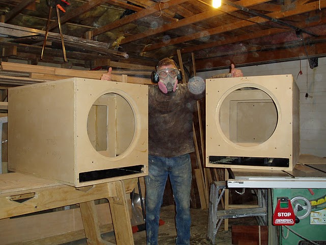 Person holding up two large plywood speaker boxes in a basement shop