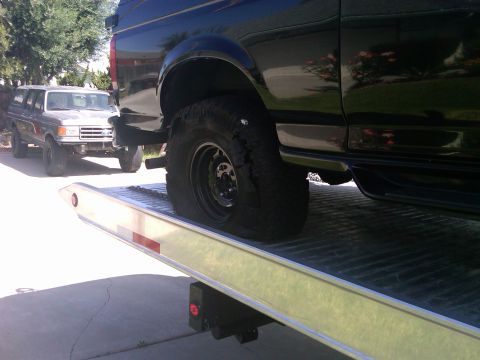 1986 Ford Bronco - A black SUV with a flat tire is loaded onto a tow truck, with an older model SUV visible in the background.