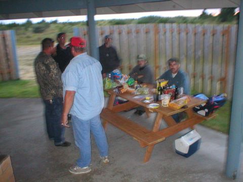 1978 Ford Bronco - Several men are socializing around a wooden picnic table laden with food and beverages, under a covered area.