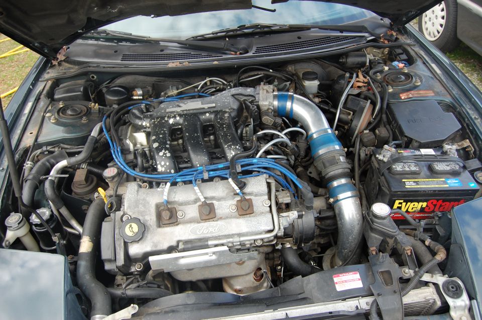 Engine bay with Ford V6 engine, blue spark plug wires, and aftermarket intake. Battery is visible