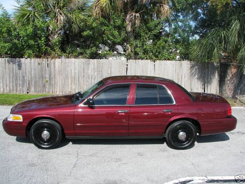 2005 Ford Crown Victoria - A maroon Ford Crown Victoria is parked on a street with a wooden fence and trees in the background.