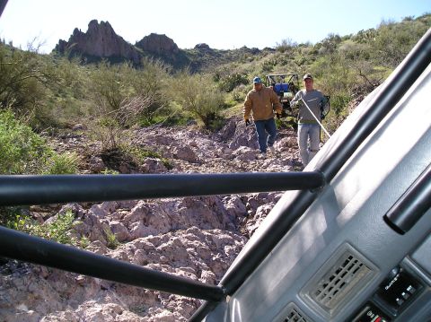 1986 Toyota Hilux - Driver's-eye view out over a gray dash and padded black roll cage bars at a rocky desert trail, with two spotters walking down the ledge to…