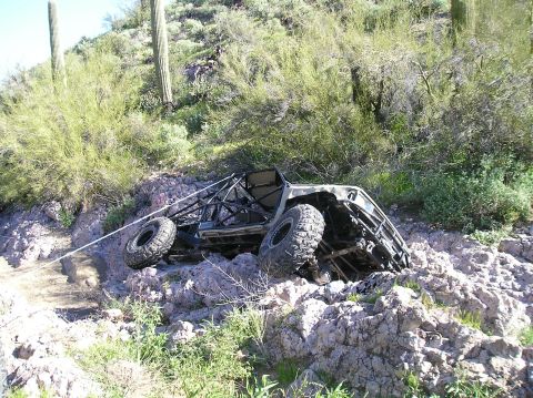 1986 Toyota Hilux - A stripped tan tube-frame rock crawler rests on its side in a boulder-strewn desert wash, its black roll cage, exposed underbody and mud-te…