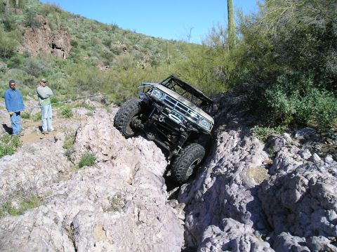 1986 Toyota Hilux - A tan chopped-top 4x4 pickup with a caged cab, winch bumper and solid front axle descends nose-down into a rock crevice on a Sonoran desert…