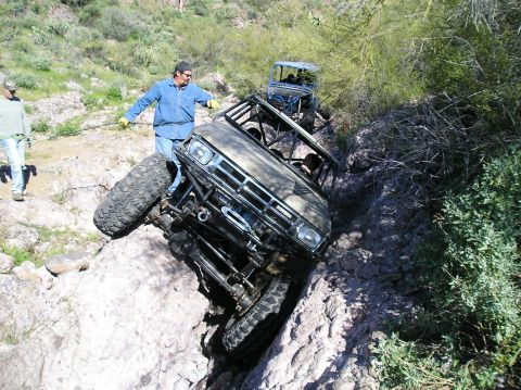1986 Toyota Hilux - Overhead front view of a tan chopped Toyota 4x4 pickup with exposed roll cage, winch bumper and TOYOTA-lettered grille dropping into a rock…
