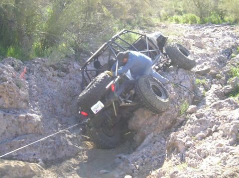 1986 Toyota Hilux - Rear three-quarter view of a caged tube-frame buggy on mud-terrain tires tipped hard against a rock ledge in a desert wash, a winch cable r…