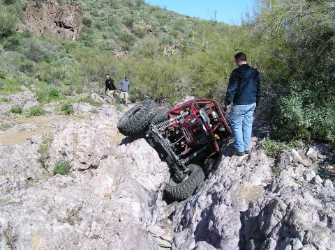1986 Toyota Hilux - A red tube-chassis rock buggy lies on its side wedged in a rocky desert wash, exposing its coilover shocks, solid axle and mud-terrain tire…