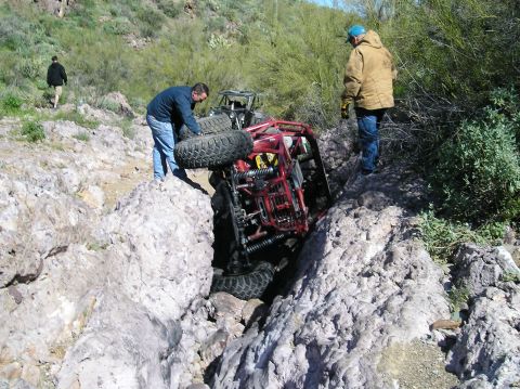1986 Toyota Hilux - A red tube-chassis rock buggy on its side wedged in a narrow rock crack on a desert trail, exposing its coilover shocks, links and axles wh…