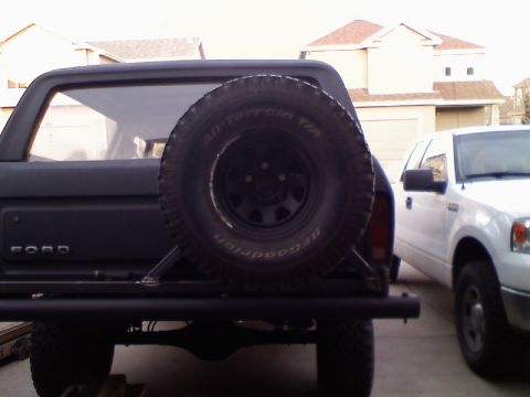 1979 Ford Bronco - Rear view of a black SUV showing its external tire carrier and a BFGoodrich All-Terrain tire mounted. A white pickup truck is visible to th…