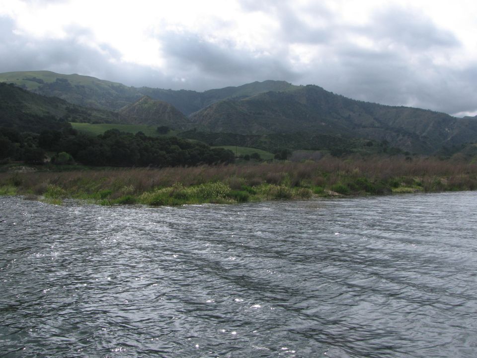 Water ripples in foreground with grassy bank and rolling hills under a cloudy sky