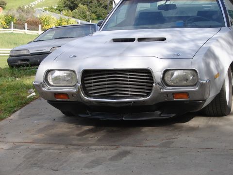 1973 Ford Ranchero - A silver pickup truck is parked in front of another vehicle, showing its distinctive grille and headlights.