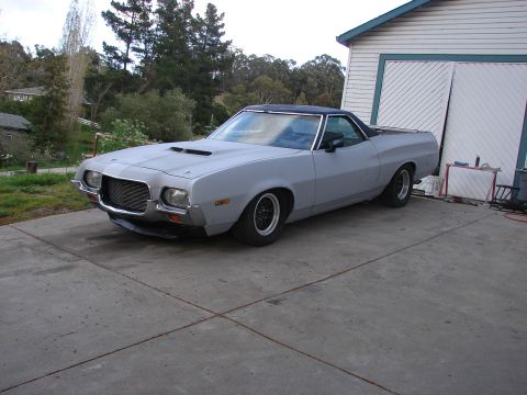 1973 Ford Ranchero - A gray-primered Ford Ranchero car-pickup with a black vinyl roof sits on a concrete pad outside a shop building, shown from the front three…