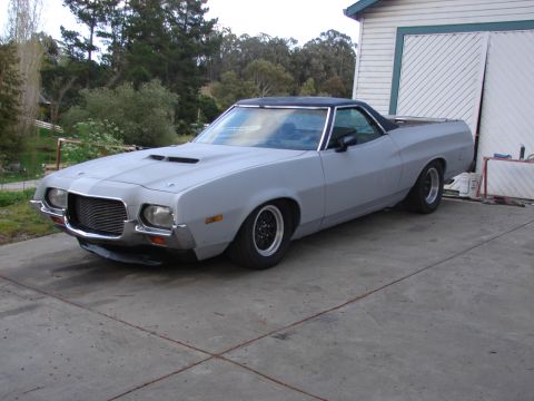 1973 Ford Ranchero - A Ford Ranchero coupe-utility in bare gray primer parked on a concrete pad beside a white garage, shot from the driver's-side front three-q…