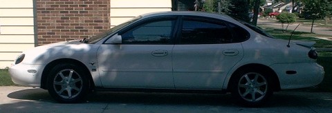 1997 Ford Taurus - Side profile of a white 1997 Ford Taurus sedan parked in front of a brick and siding house.