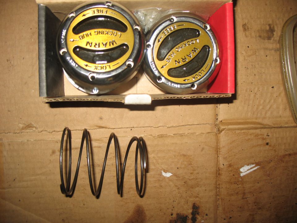 Two chrome and yellow Warn locking hubs in a box with a metal spring on cardboard