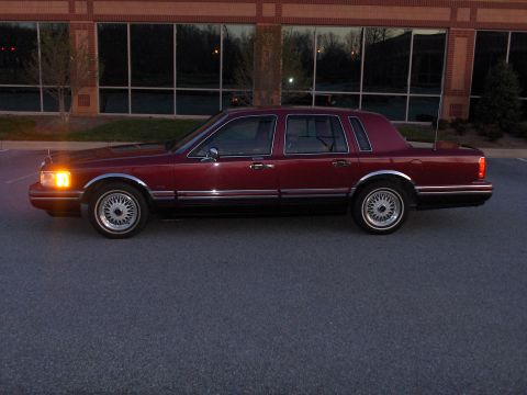 1993 Lincoln Town Car - Side profile of a maroon 1993 Lincoln Town Car parked in front of a brick building with large windows.