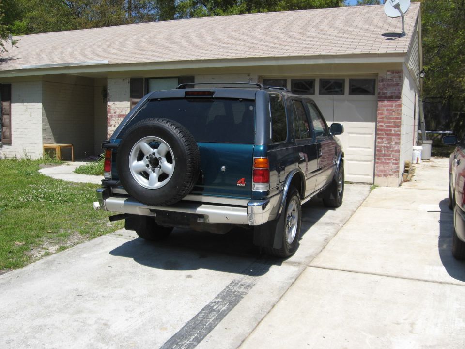 Teal SUV parked in driveway, rear view. Spare tire on back, chrome bumper, mud flaps. House and garage in background