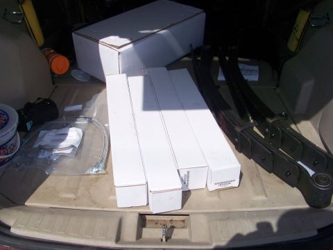1989 Ford Bronco - Suspension parts laid out on a tan plastic cargo/tailgate liner: four long white shock absorber boxes, a larger white parts carton, a bagge…