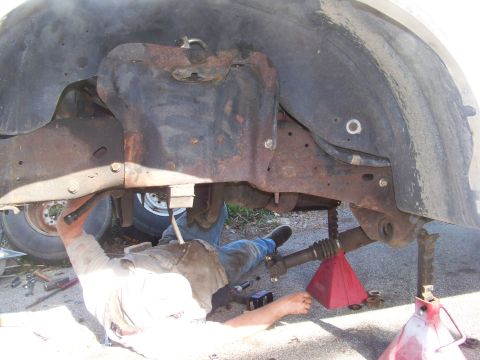 1989 Ford Bronco - Underbody view of a truck up on red jack stands in a driveway, showing the surface-rusted frame rails, crossmember and transmission crossme…