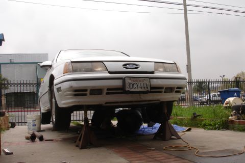 1989 Ford Taurus - A white 1989 Ford Taurus is raised on jack stands, with a mechanic visible underneath.