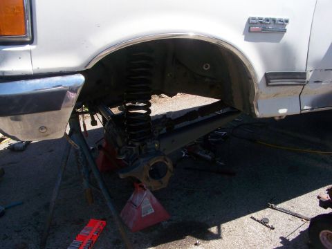 1989 Ford Bronco - Front axle swap in progress on a white Ford Bronco: the wheel and hub are off, exposing the black coil spring, shock and the bare Dana knuc…