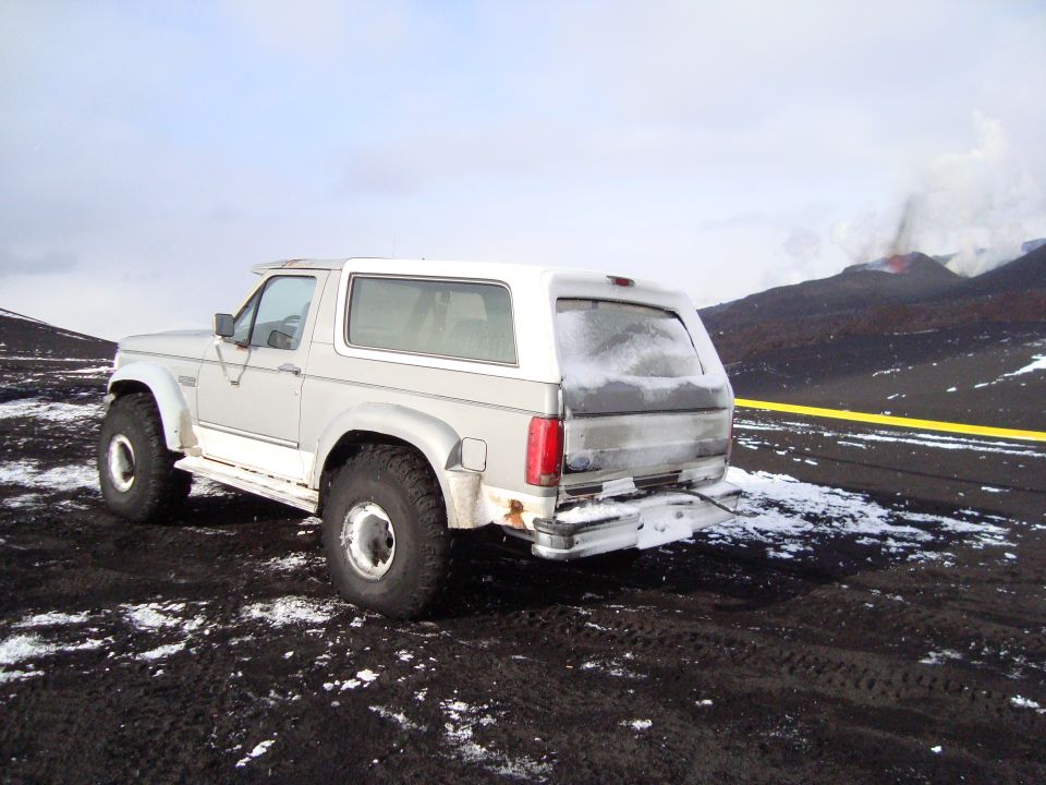 1992 Ford Bronco parked on dark ground with snow, volcano erupting in background