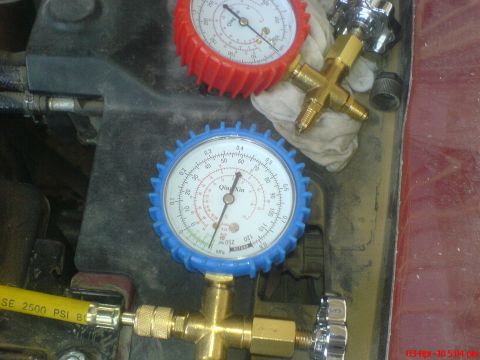 1998 Ford Crown Victoria - Overhead view of a Qing Xin A/C manifold gauge set resting on a dark engine bay panel, with the blue low-side gauge needle pulled into vacu…