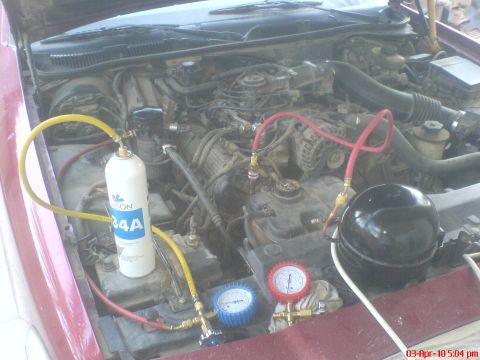 1998 Ford Crown Victoria - Engine bay of a maroon/red sedan with an A/C recharge setup connected, including a can of R-134A refrigerant, yellow and red charging hoses…
