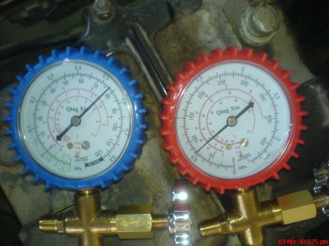 1998 Ford Crown Victoria - Close-up of the blue low-side and red high-side Qing Xin A/C manifold gauges lying on a rusty engine bracket, both needles reading roughly…
