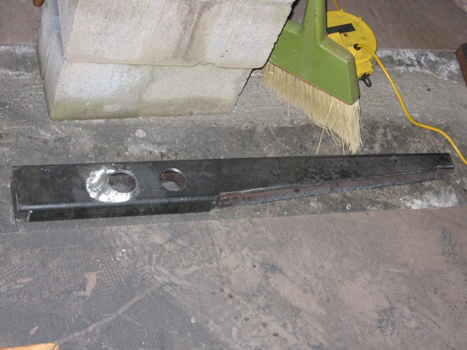 Dark metal automotive part with two circular holes and welded seams, lying on a concrete floor
