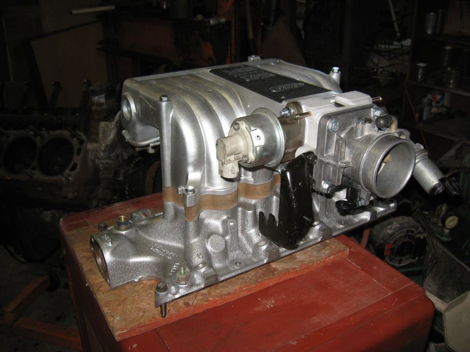 Ford intake manifold with throttle body and sensors, sitting on a red surface. Another engine block is visible in the background