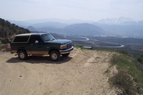1995 Ford Bronco - A green 1995 Ford Bronco with tan trim sits on a dirt road, offering a view of a winding road and distant mountains.