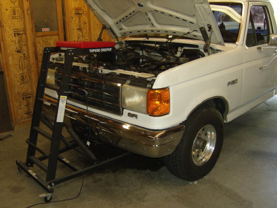 White Ford F-150 truck with hood open, parked in a garage with a topside creeper positioned in front