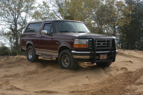 1996 Ford Bronco - A maroon Ford Bronco with tan trim is shown driving on a sandy terrain.