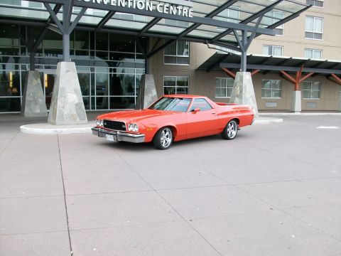 1973 Ford Ranchero - Red Ford Ranchero coupe utility parked alone on the concrete forecourt of a convention centre, framed by stone-clad pylons and a glass entr…